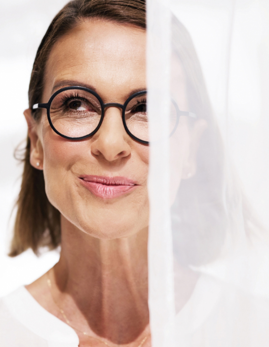 Frau im Gleitsichtalter trägt Brille uns schaut hinter Vorhang hervor.